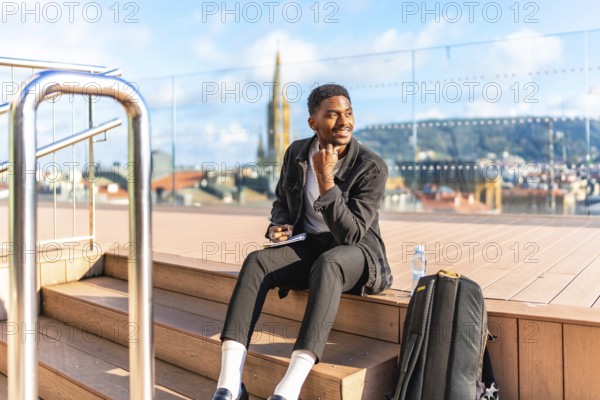 Happy african man sketching by hand in a notebook sitting on a rooftop with city views