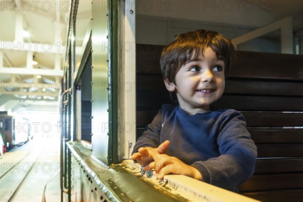 A young boy is sitting on a bench in a train station, looking out the window with a smile on his face