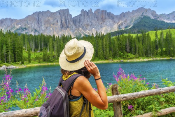 Woman with straw hat and backpack admiring the turquoise waters of lake carezza with the latemar massif in the background, dolomites, italy