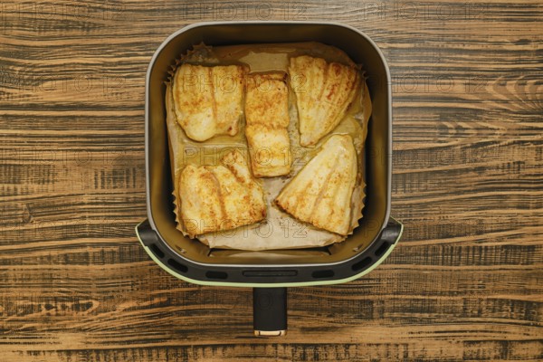 Five golden brown grilled fish fillets are neatly placed inside an air fryer basket. The background features a rustic wooden table, showcasing a cozy cooking environment