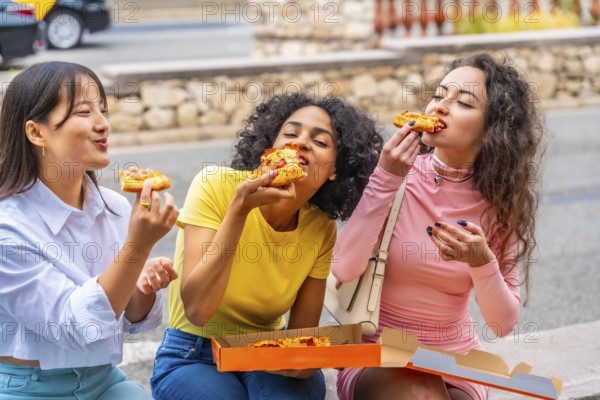 Hungry female multiracial friends eating take away pizza sitting outdoors in the city
