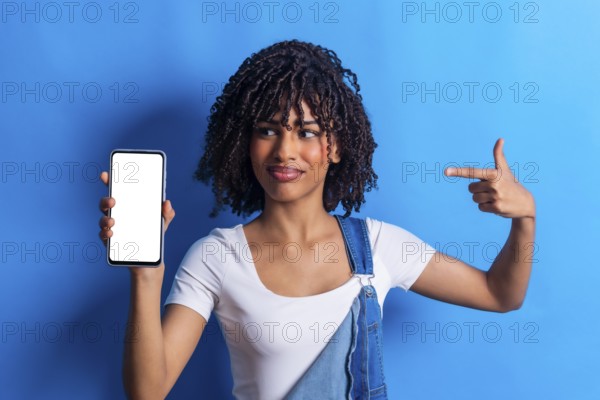 Studio portrait of a young woman holding a smartphone with blank white screen and pointing at it with index finger, isolated on a blue background