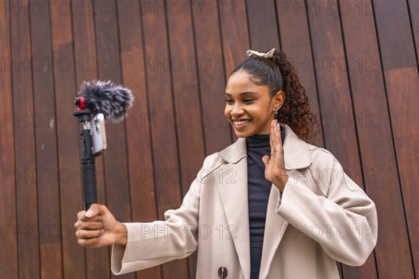 Young african american woman influencer waving at camera while recording vlog outdoors next to a wooden facade in the city