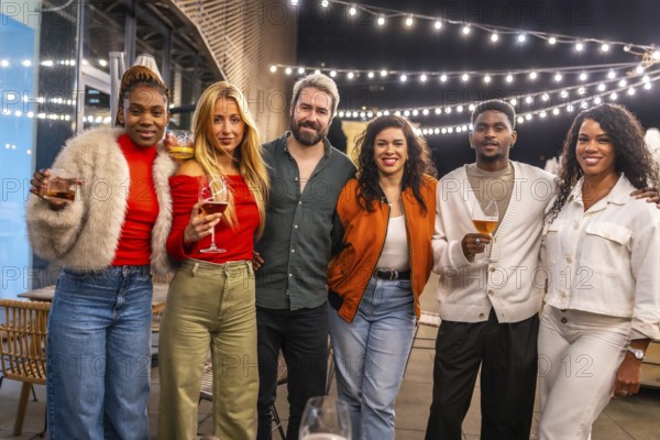 Portrait of a multi-ethnic group of people enjoying at bar terrace standing with drinking glasses smiling at camera together at night