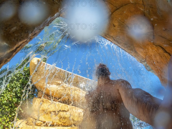 Man refreshing under a waterfall in a water park during summer vacation, enjoying the cool water on a hot day