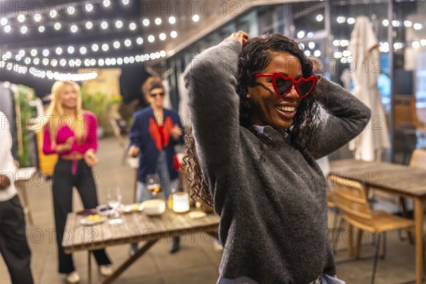 Funny latin woman with heart-shaped sunglasses and casual winter clothes dancing next to friends at night party in a roof bar terrace
