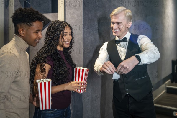 Young couple holding popcorn buckets entering a movie theater and checking their tickets with the usher before watching a film