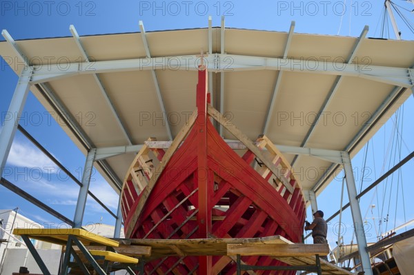 Front view of a partially built red wooden ship in a covered shipyard under a clear sky, Patmos Marine, boatyard, Diakofti, Patmos, Dodecanese, Greek Islands, Greece, Europe