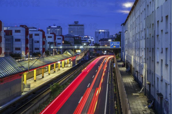Essen, North Rhine-Westphalia, Germany, A40 motorway in the city centre at dusk. Deserted Savignystrasse underground station. On the right, residential buildings behind a noise barrier. On the wall is a LANUV air monitoring station, Essen Kruppstrasse 117 (EKRU2), on the right under the lantern. Residents are affected by noise and exhaust fumes. At the back, buildings with Postbank, Eon and Evonik lettering. Moon in the sky, Europe