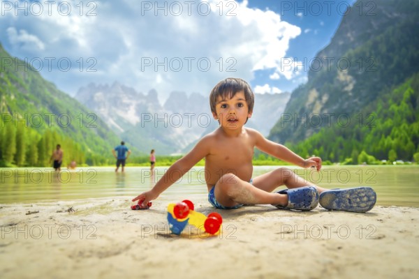 Young boy playing with toy cars on the sandy shore of lago di landro, surrounded by the stunning peaks of the dolomites in summer