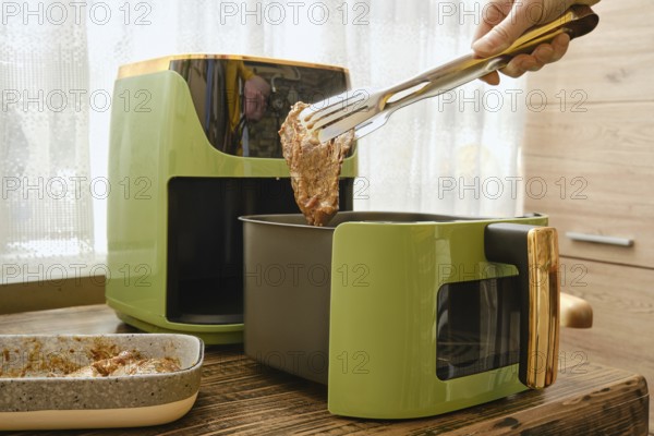 A hand holds tongs putting a piece of marinated meat to an air fryer. The process of preparing crispy and juicy meal at home
