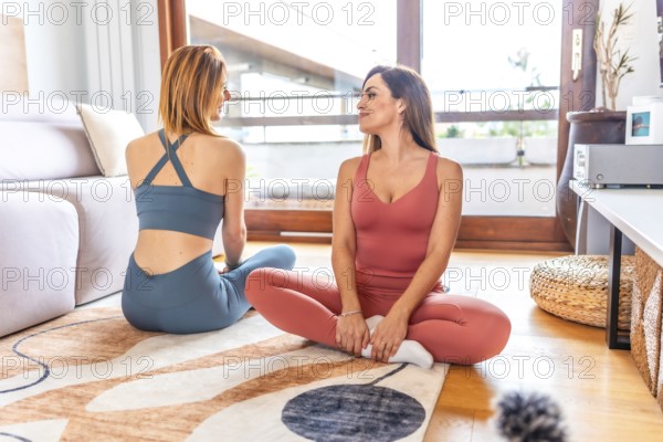 Two fitness women are sitting on a rug in a living room, enjoying a relaxing moment during their online yoga class