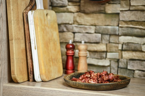 A rustic kitchen features a plate of chopped bacon next to wooden cutting boards. Two shakers, one wooden and one red, sit in the background, adding to the warm atmosphere