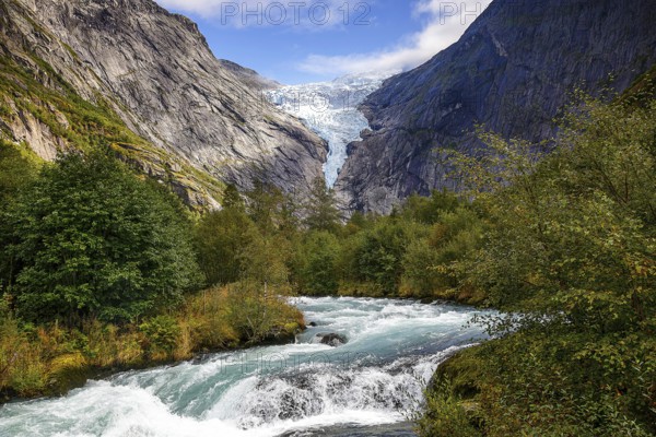 River flanked by mountains flows through a green valley with a view of the shimmering Briksdalsbreen glacier in Norway