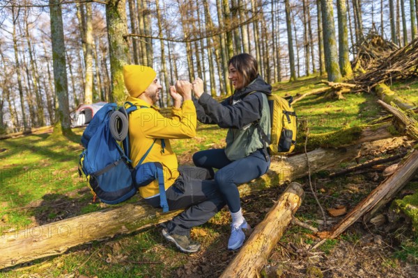 Hikers with backpacks are sitting on a fallen tree trunk in a forest, giving each other a high five while resting during their camping trip