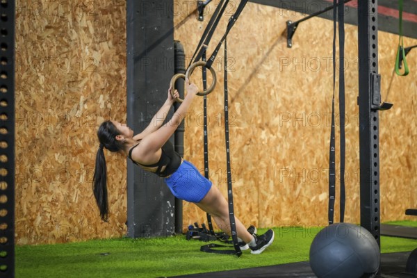 Full length side view of an asian strong woman exercising biceps using gym rings in a modern gym