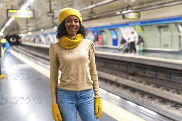 Smiling young woman wearing yellow winter clothes waiting for subway train arrival at station platform