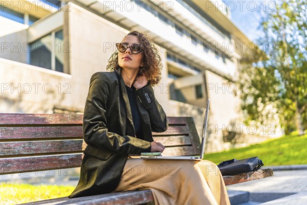 Young arab female freelancer working with laptop sitting on park bench under the sun