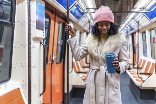 Smiling young black woman holding reusable coffee cup while standing on moving subway train