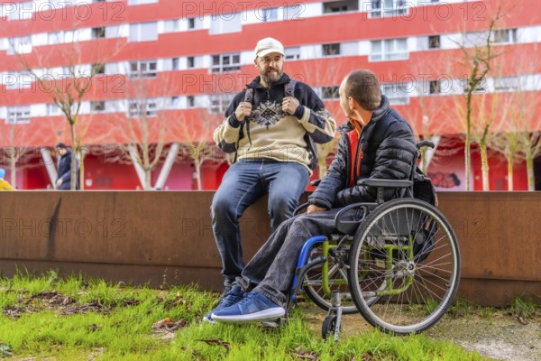 Two male friends, one using a wheelchair, are sitting on a low wall, chatting and enjoying their time together in an urban setting