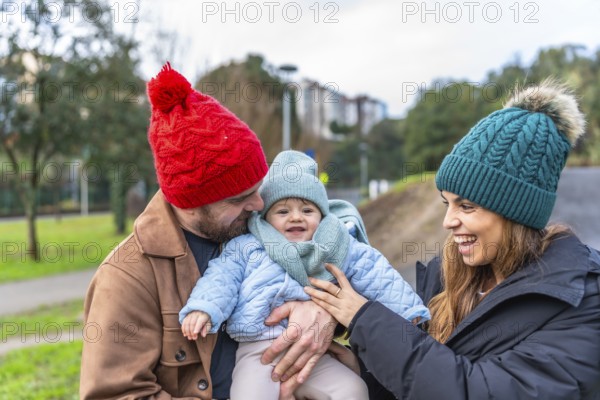 Parents holding their baby while smiling and enjoying a joyful winter day in a park, surrounded by nature and the beauty of the season