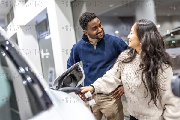 Smiling couple examining the charging socket of an electric vehicle while exploring options at a car dealership, embracing sustainable living