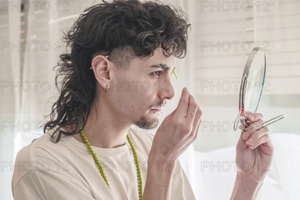 Concentrated young man with curly hair plucking his eyebrows with tweezers while looking in a small mirror, taking care of his appearance in a bright room