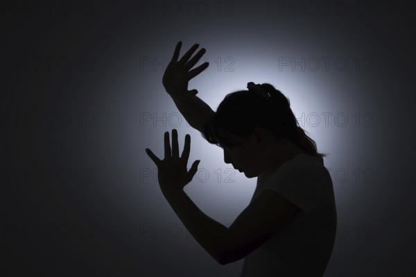 Symbolic photo on the subject of violence against woman. The silhouette of a young woman with her hands held protectively in front of her face is silhouetted against a white wall. Berlin, 22 August 2024
