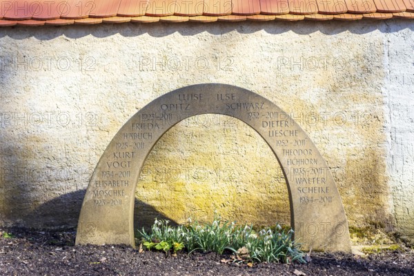 Gravestone in the shape of a rainbow on a communal urn grave, with snowdrops (galanthus) blooming in front of it, Merzdorf cemetery, Riesa, Saxony, . Germany