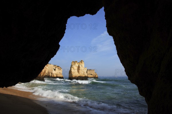 Praia do Pinhao, Lagos, Algarve, Portugal, View from a cave to the sea with waves, a sandy beach and rocks in the background under a blue sky, Europe