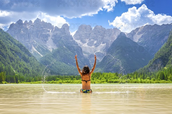 Young woman raising arms and splashing water in the famous lago di landro, also known as durrensee lake, in dolomites, italy, during summer vacation