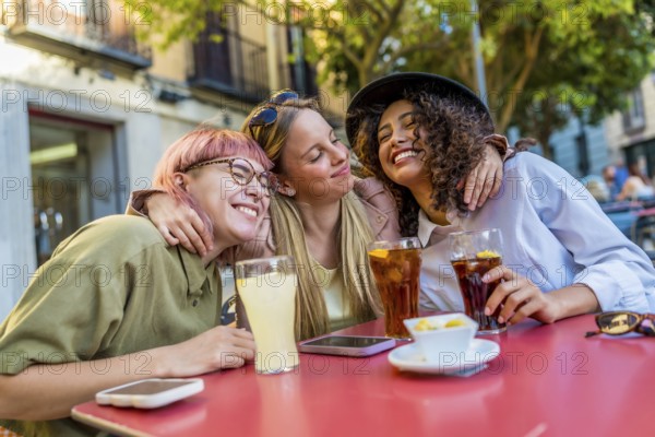 Diverse female friends embracing and smiling sitting on the terrace of a bar