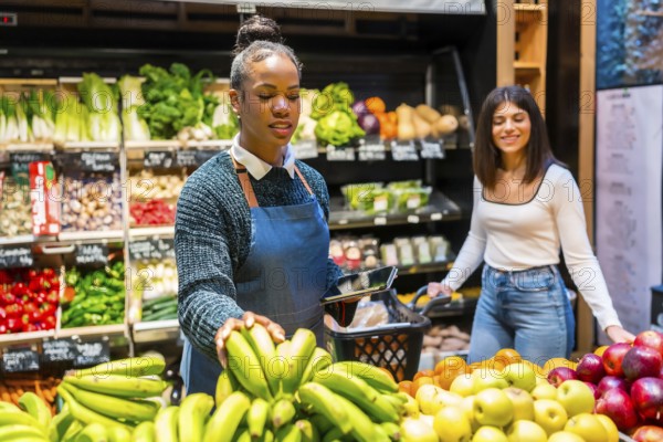 Greengrocer arranging fresh bananas while using digital tablet and helping customer in organic supermarket with colorful fruit and vegetables