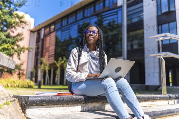 Distracted african woman looking up while using laptop sitting outside the university campus