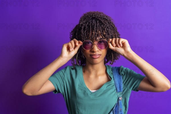 Stylish young woman lowering her purple sunglasses while posing in a vibrant purple studio, creating a trendy and fashionable image