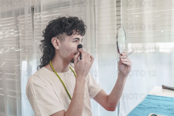 Young man with curly hair is applying makeup to his face with a brush. While holding a small mirror in his other hand. Showcasing self-expression and challenging traditional gender roles in beauty