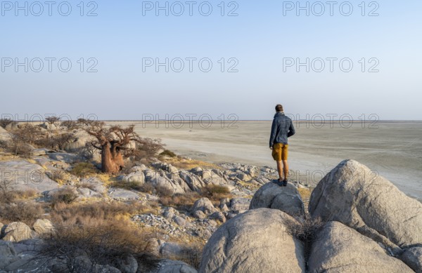 Tourist looking over the salt pan, African baobab or monkey bread tree (Adansonia digitata), between round rocks, at sunrise, with salt pan, Kubu Island (Lekubu), Sowa Pan, Makgadikgadi salt pans, Botswana, Africa