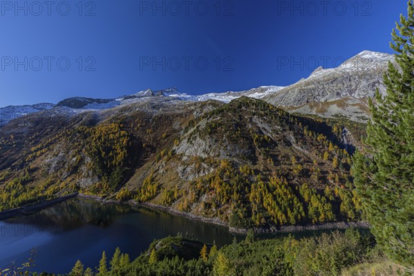 The Galgenbichl reservoir with a mountain forest in fall foliage on a bright sunny morning. Seen from the Malta Hochalm road. Carithia, Austria, Europe