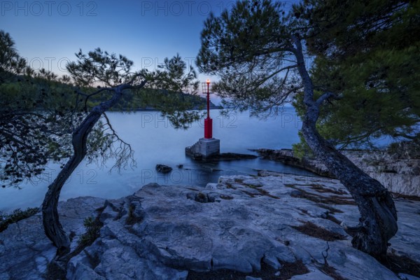 Lighthouse red sea mark between trees on rocks on a calm sea at dusk, Hvar, Croatia, Europe