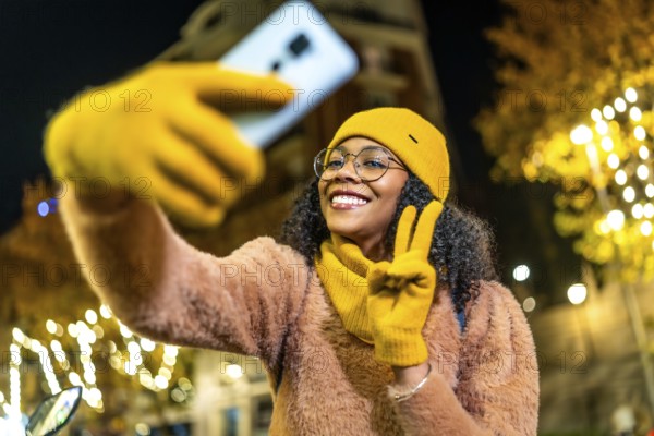 Smiling woman wearing yellow winter clothes taking selfie with smartphone and making peace sign in christmas decorated city street at night