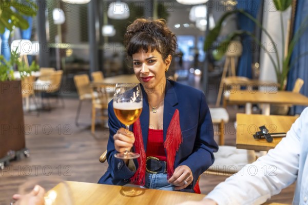 Modern woman toasting at camera with beer at night bar roof terrace sitting next to friends
