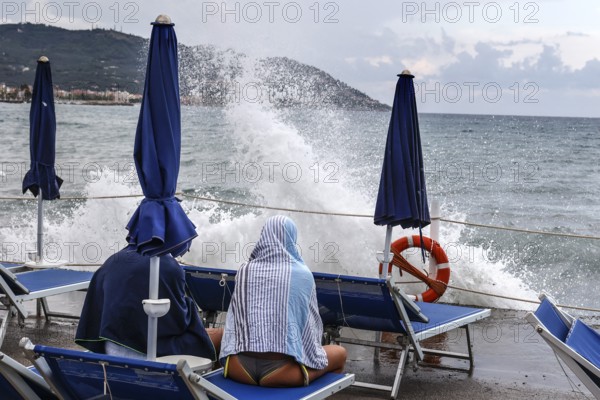 Waves crashing against the pier of a bathing jetty where a couple is sitting on deckchairs, Diana Marina, Italy, 18/08/2024, Diano Marina, Liguria, Italy, Europe