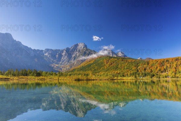 Lake Almsee in autumn with a forest reflected in the lake. A slowly dissipating fog moves over the lake. Blue sky with no clouds