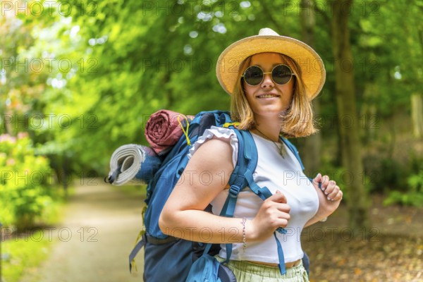 Cheerful caucasian young woman wearing sunglasses and sun hat carrying backpack walking along a path enjoying nature in the forest