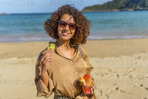 Portrait of a beaut cyute young arab woman with curly hair and sunglasses eating sliced tropical fruit at beach
