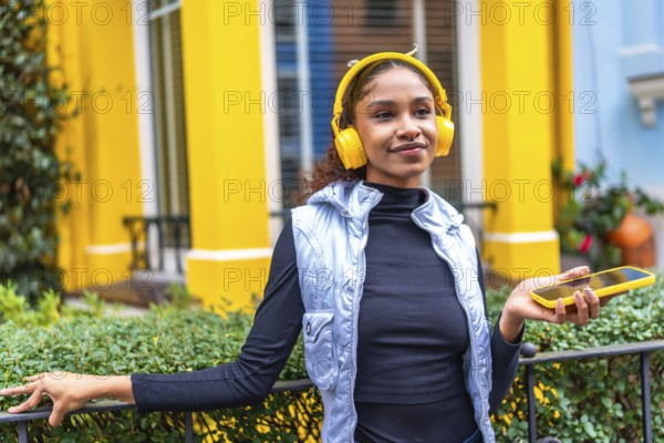 Afro woman listening to music relaxes in the city leaning on garden rail of a colorful yellow house