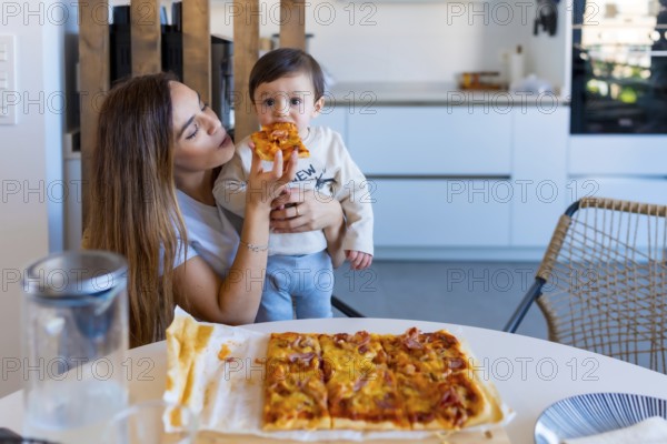 Mother enjoying a moment of togetherness while feeding her baby a slice of pizza at the kitchen table during a cozy family meal