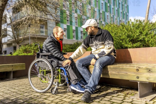 Two friends talking and smiling on a bench in a city park, one of them is using a wheelchair