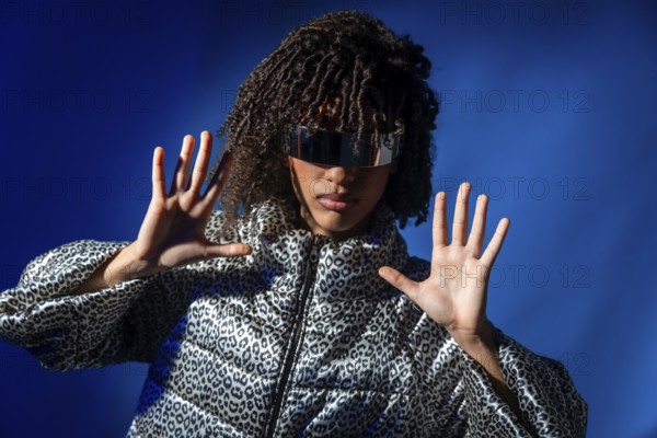 Stylish model wearing futuristic visor and patterned jacket pushing with hands on vibrant blue background, showcasing modern fashion and technology
