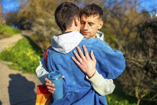 Two young men hug tightly in a park, expressing their love and affection, with a pride flag visible